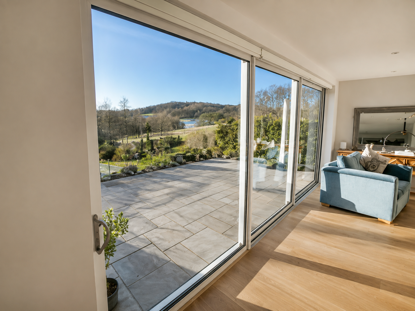 White aluminium sliding patio doors opening onto a large paved terrace with landscaped gardens and countryside views, creating a bright and spacious living area