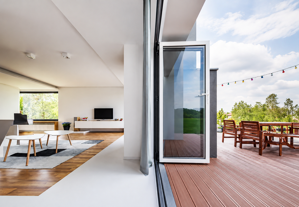 Modern aluminium bifold doors fully opened, creating a seamless connection between a bright contemporary living room and a timber decked outdoor dining area.