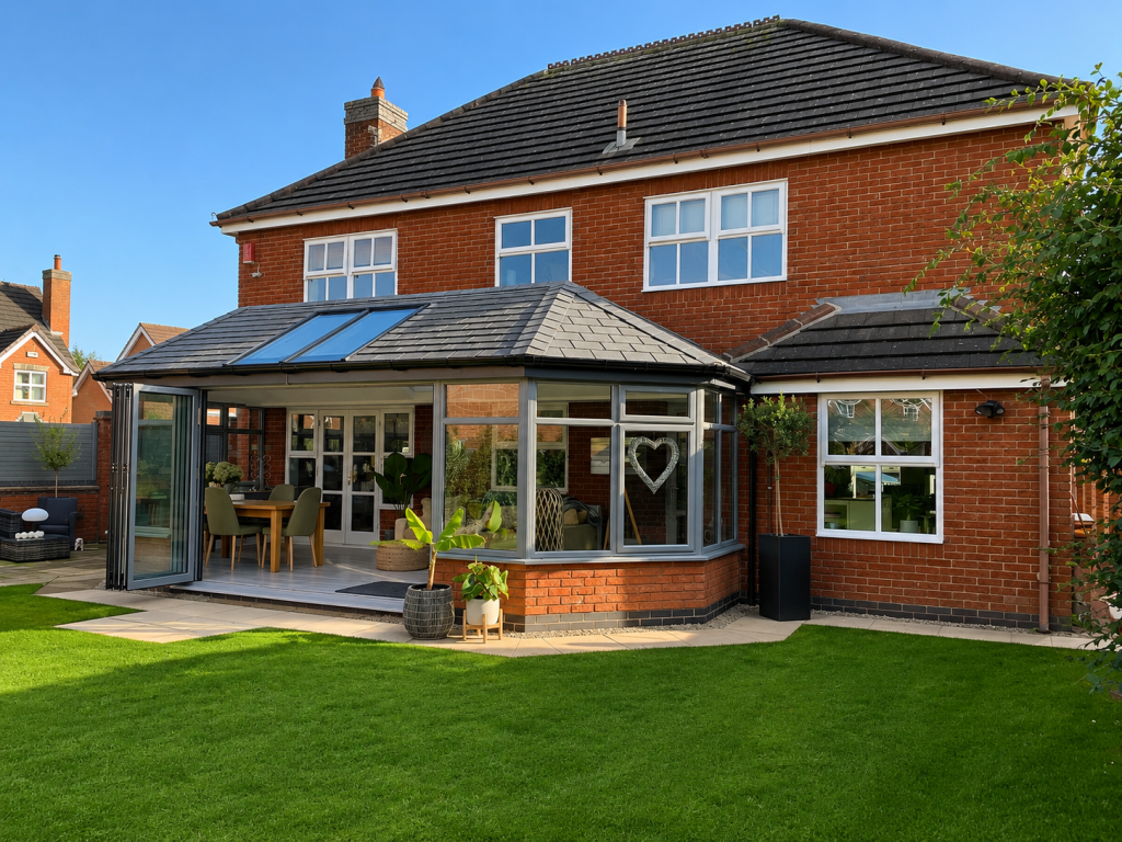 Modern aluminium garden room extension with anthracite grey frames, bi-fold doors, roof lantern glazing and a tiled roof, installed on a detached red brick home overlooking a landscaped garden.