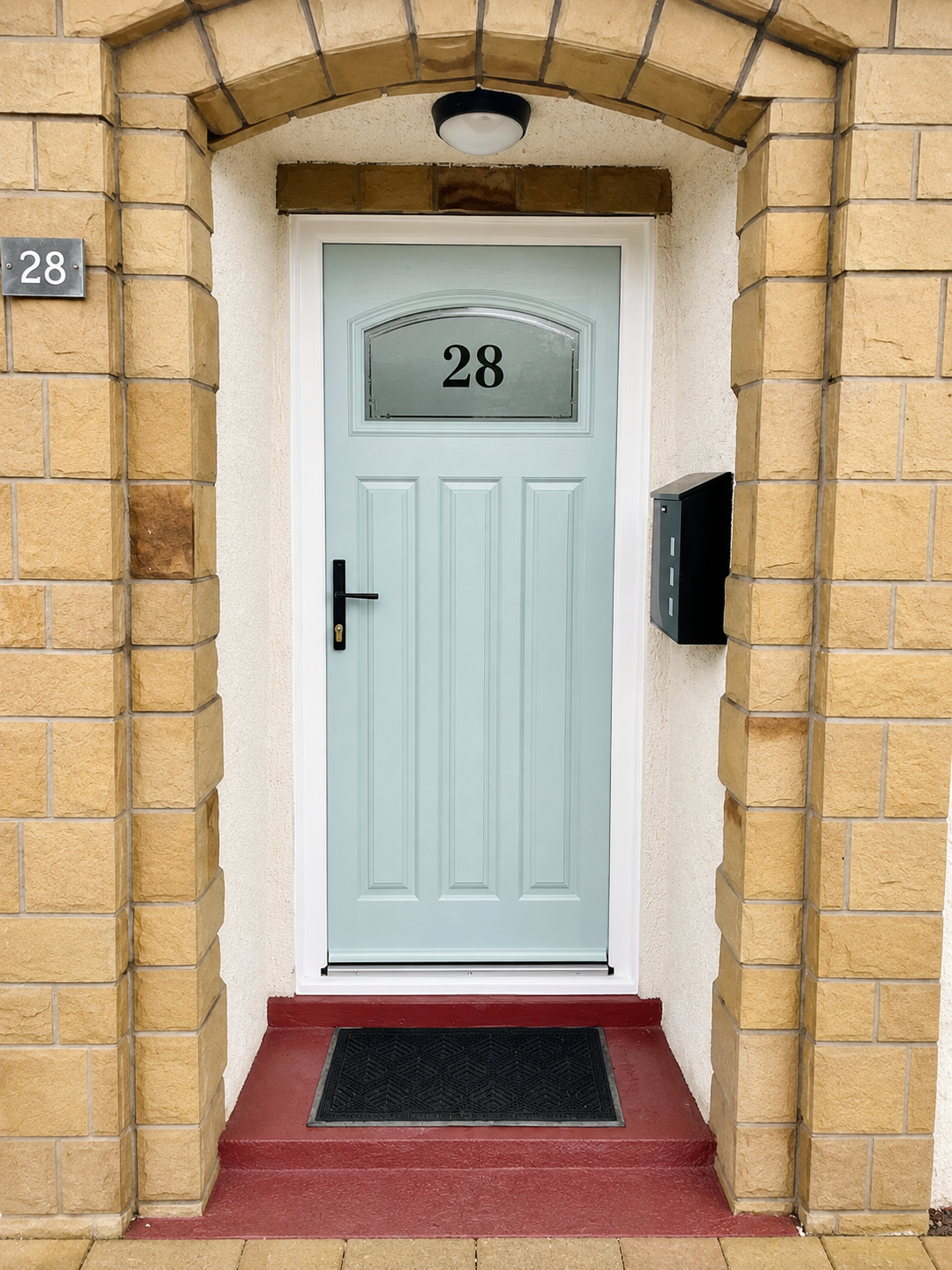 Light blue composite entrance door with an arched glazed panel, black hardware, and white frame, installed beneath a stone archway at the entrance to a modern home.