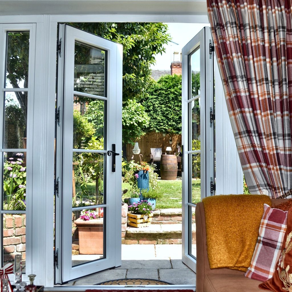 White French doors opening onto a sunny garden, with brick steps, plants and outdoor seating beyond.